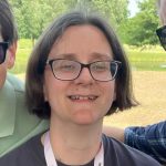 An image of Halycopter. She's standing outside with greenery in the background. She's smiling broadly and has brown hair and is wearing glasses. You can see a hint of her Halycopter t-shirt and is wearing a lanyard.