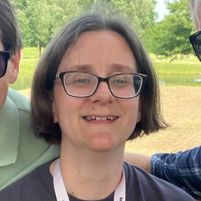 An image of Halycopter. She's standing outside with greenery in the background. She's smiling broadly and has brown hair and is wearing glasses. You can see a hint of her Halycopter t-shirt and is wearing a lanyard.